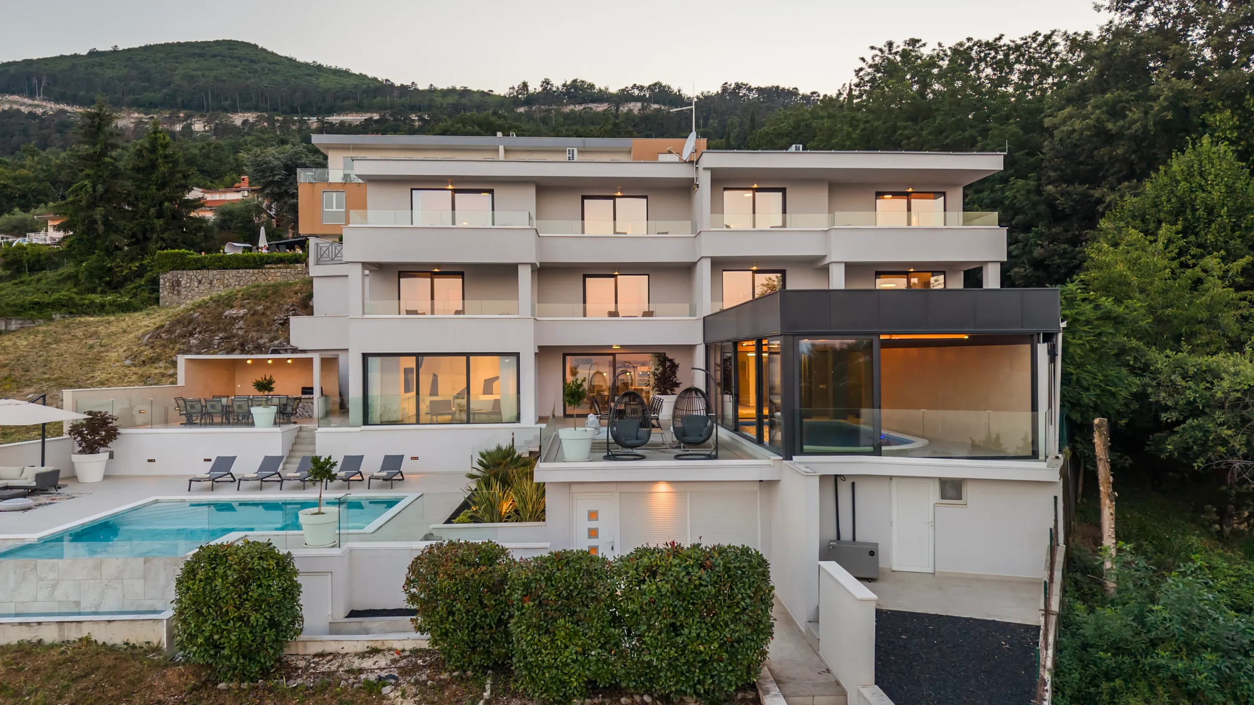Villa Theodor's terrace with egg chairs and glass-enclosed indoor pool at dusk overlooking the hillside village
