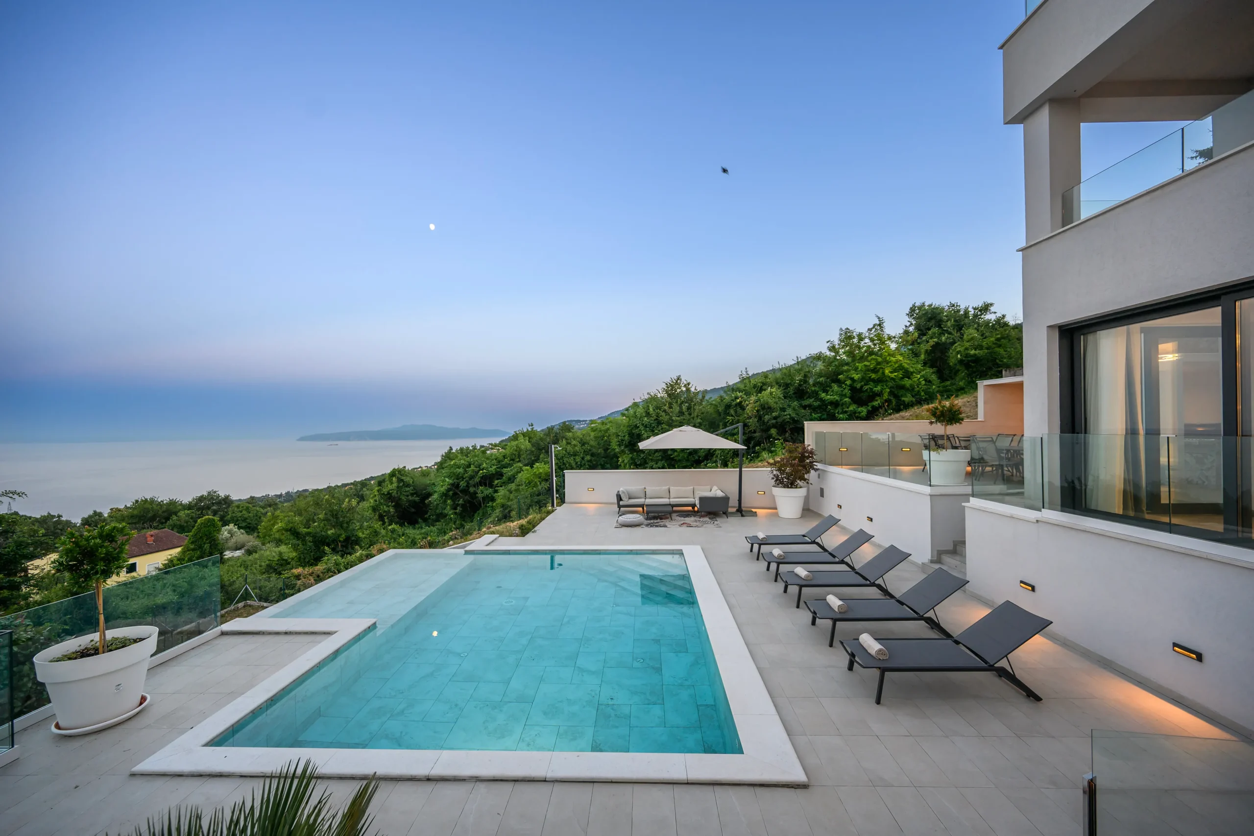 Infinity pool and terrace of Villa Theodor at dusk with the moon rising over the Kvarner Bay